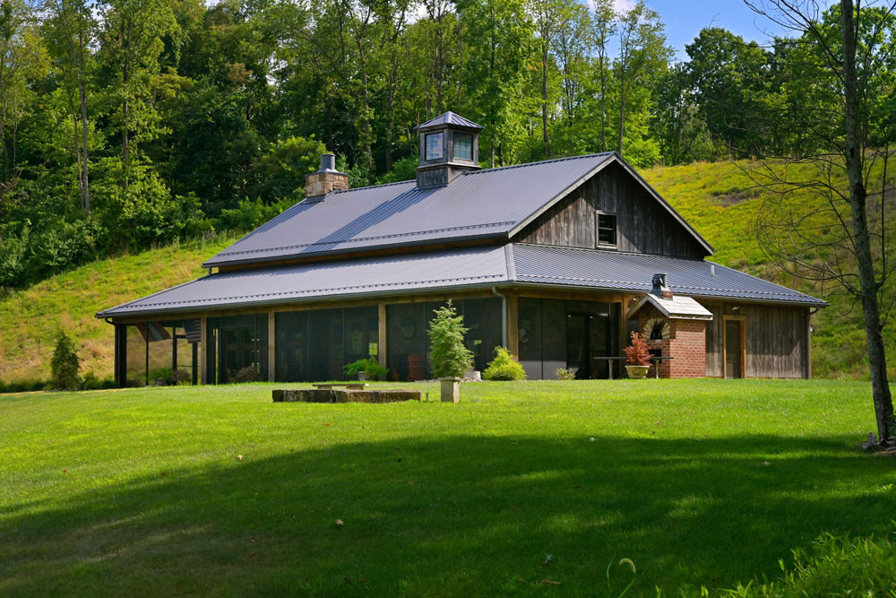Custom-built party barn with timber frame construction, dark wood siding, and metal roof set against a wooded hillside. Features include a stone chimney, cupola, wraparound porch, and expansive lawn—showcasing Sugarcreek Builder’s craftsmanship in designing rustic event spaces and barn-style homes.