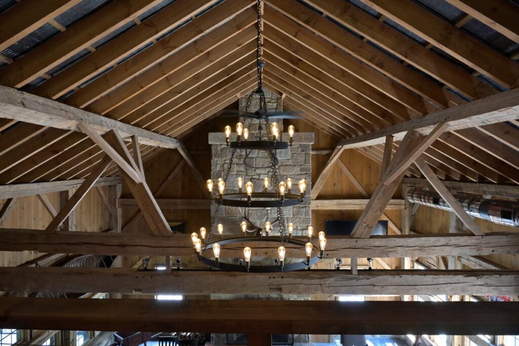 Rustic timber frame barn interior with exposed wooden trusses, vaulted ceiling, and stone fireplace. A large wrought-iron chandelier with Edison bulbs hangs prominently, showcasing Sugarcreek Builder’s craftsmanship in custom barn construction and post-and-beam design.