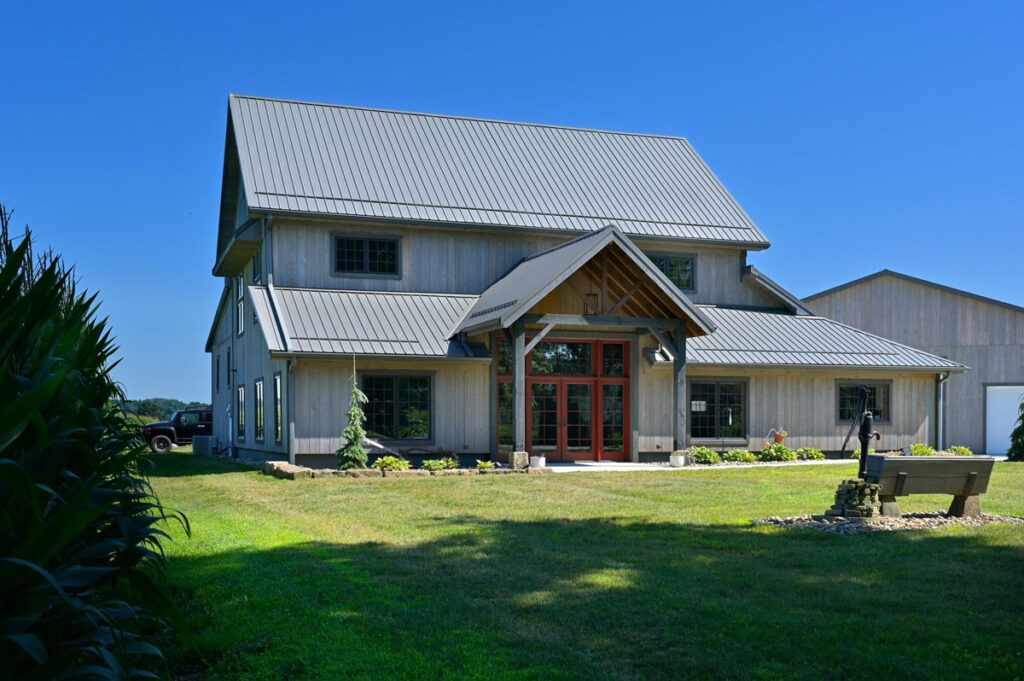 Custom timber frame barn-style home with metal roof, large front gable entry, and red-trimmed windows. Surrounded by open farmland and blue skies, this modern barn home showcases Sugarcreek Builder’s craftsmanship in post-and-beam construction and rural-inspired residential design.
